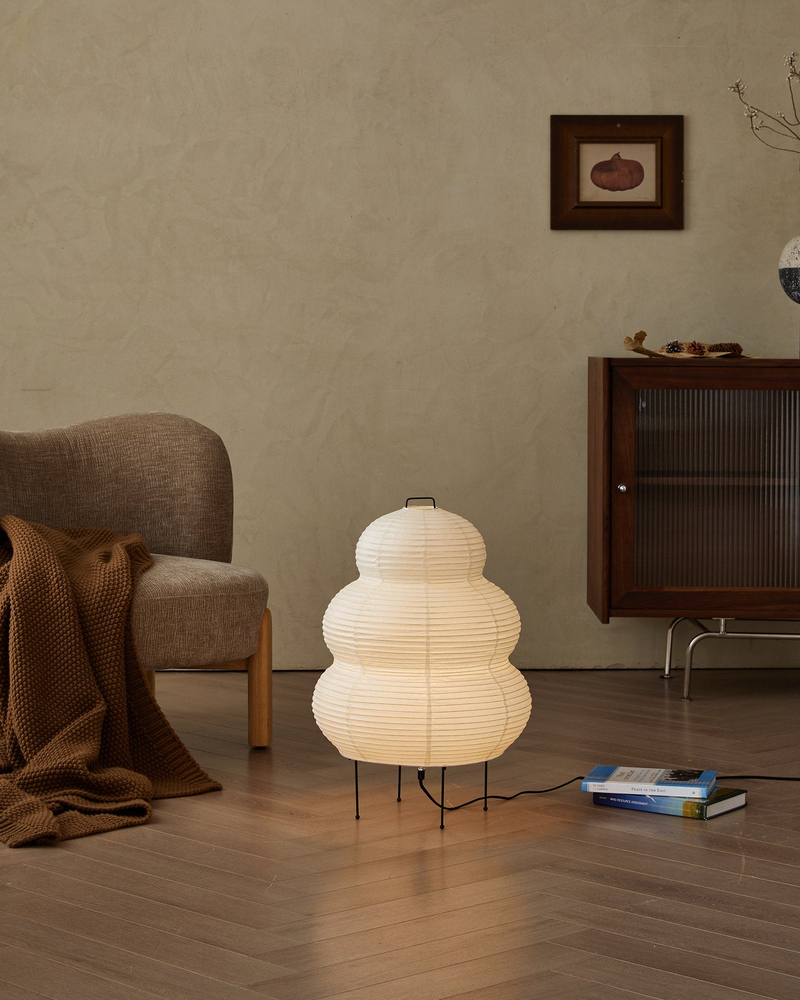 Kumo washi paper floor lamp glowing on the floor beside an armchair with a throw blanket and a book