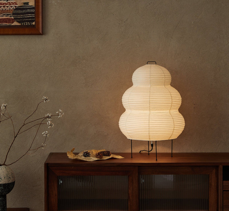 Kumo washi paper lamp lit on a walnut sideboard against a plaster wall, beside a spray of dried branches